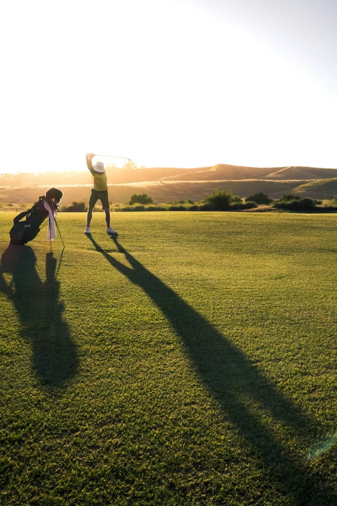 Golfer swinging golf club on grassy course at sunset, long shadows, hills in background.